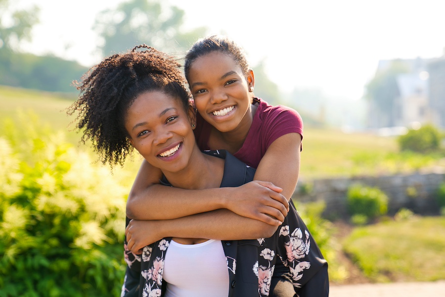 Two smiling African American sisters hug outside surrounded by green plants on a sunny day