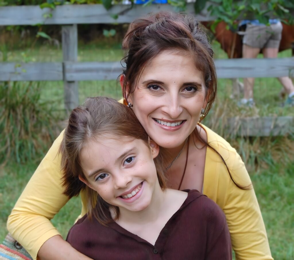 Mother and young daughter smiling close together outdoors in front of a wooden fence.