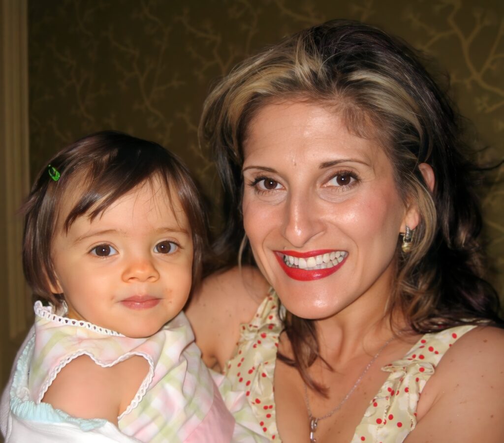 Mother holding toddler daughter indoors, both smiling close together.